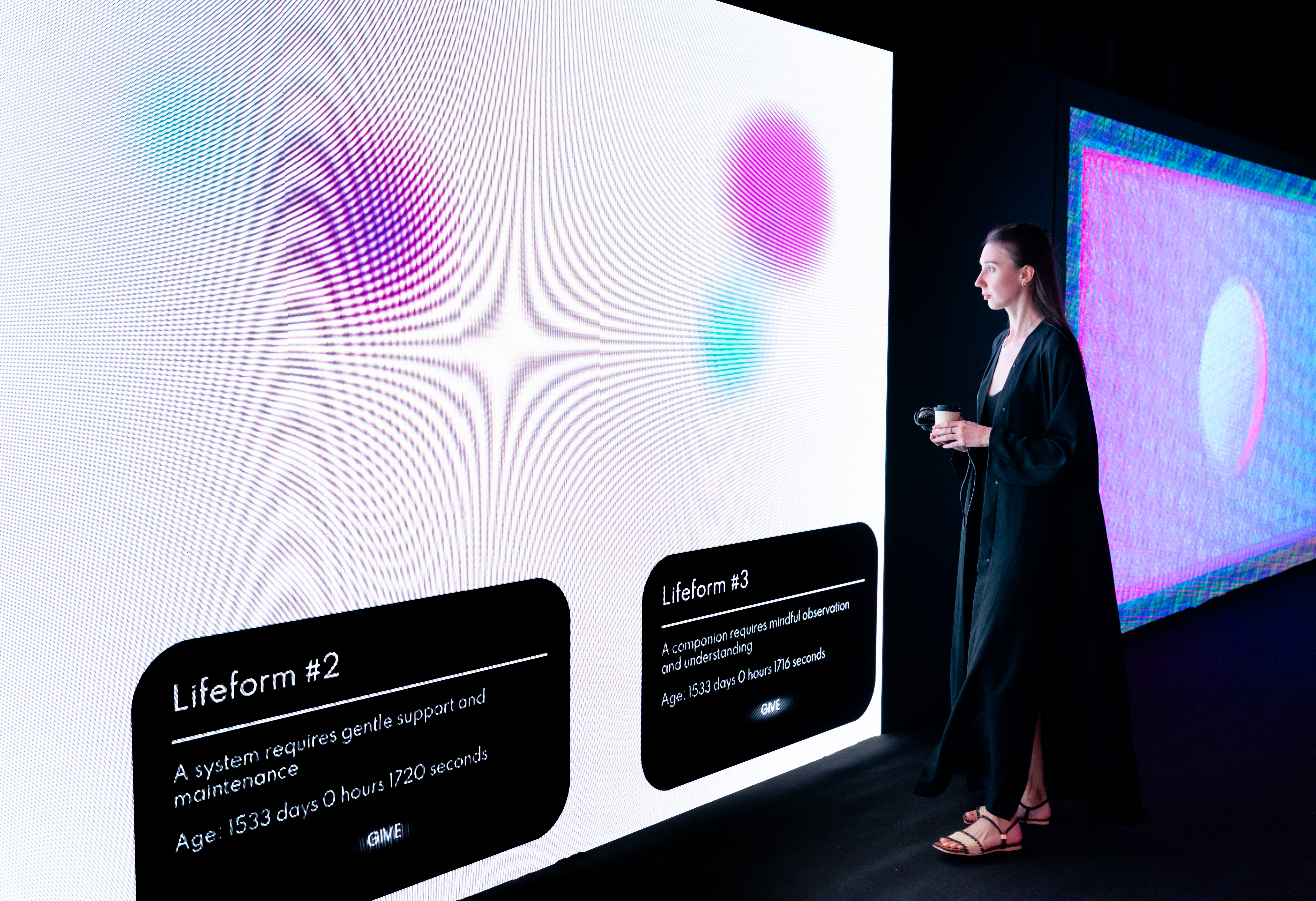 A person in a black dress stands in a darkened gallery space looking at a large bright wall display showing two Lifeforms, glowing colourful blob-like creatures, each with a description card showing their age in days and a care instruction. To the right is a second screen.