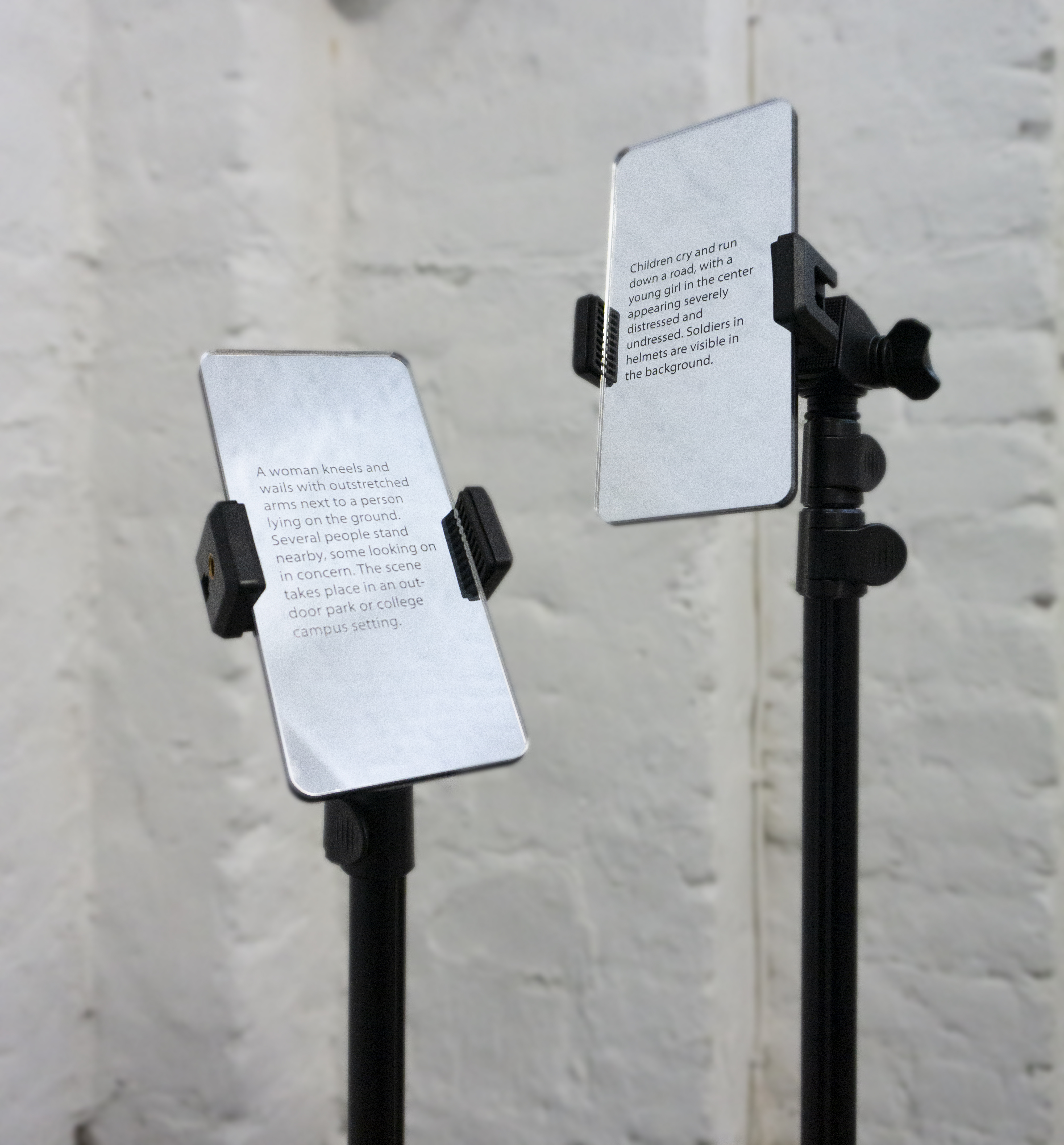 Two phone-sized mirrored metallic cards mounted on black adjustable stands against a grey wall, each displaying printed alt-text descriptions of famous photojournalism images. The left reads about a woman kneeling and wailing beside a person lying on the ground, the right about children running on a road with soldiers in the background.