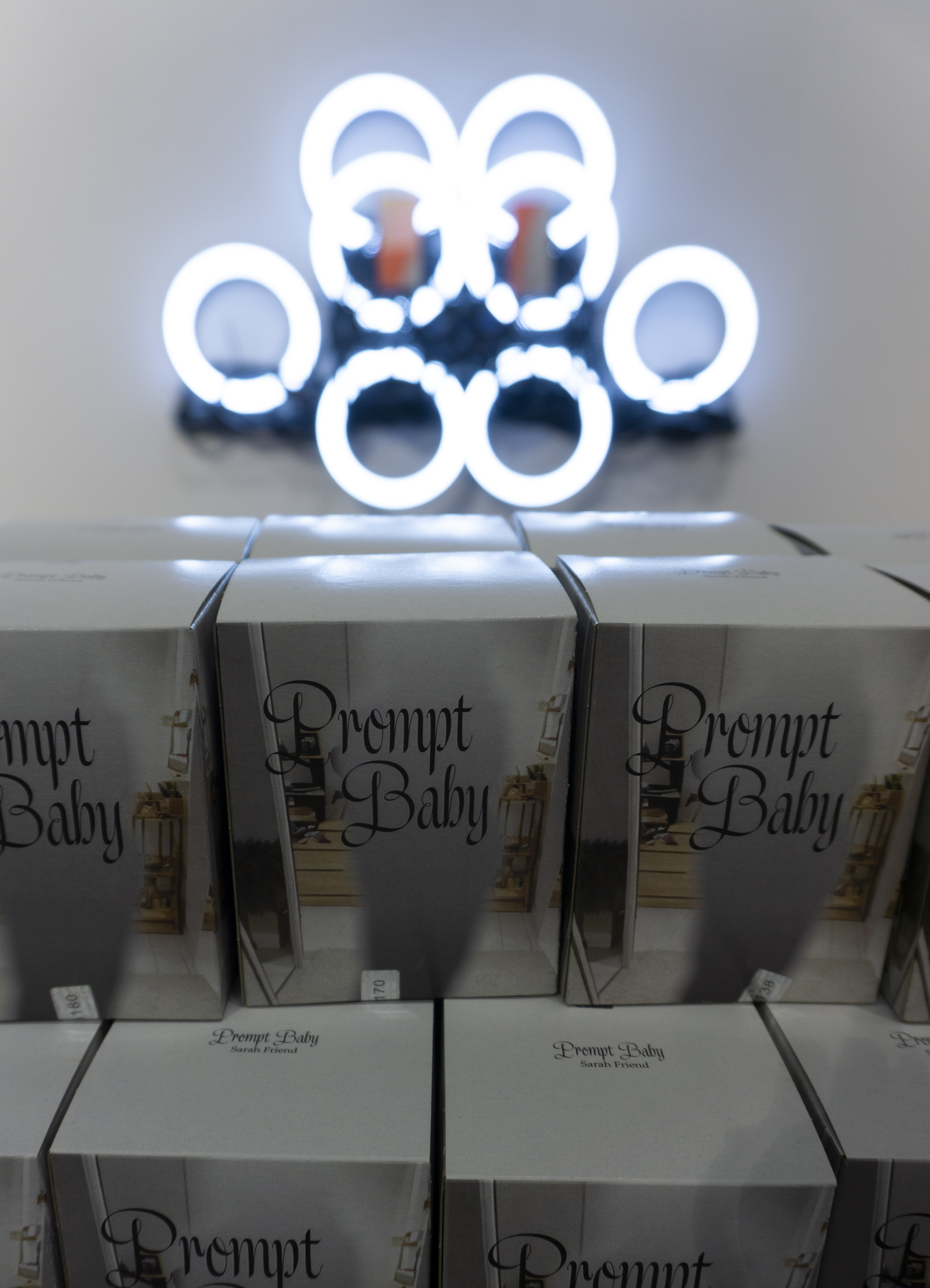 Multiple silver boxes labelled Prompt Baby by Sarah Friend stacked in a display, lit from above by a glowing sculpture made of overlapping white LED ring lights mounted behind them