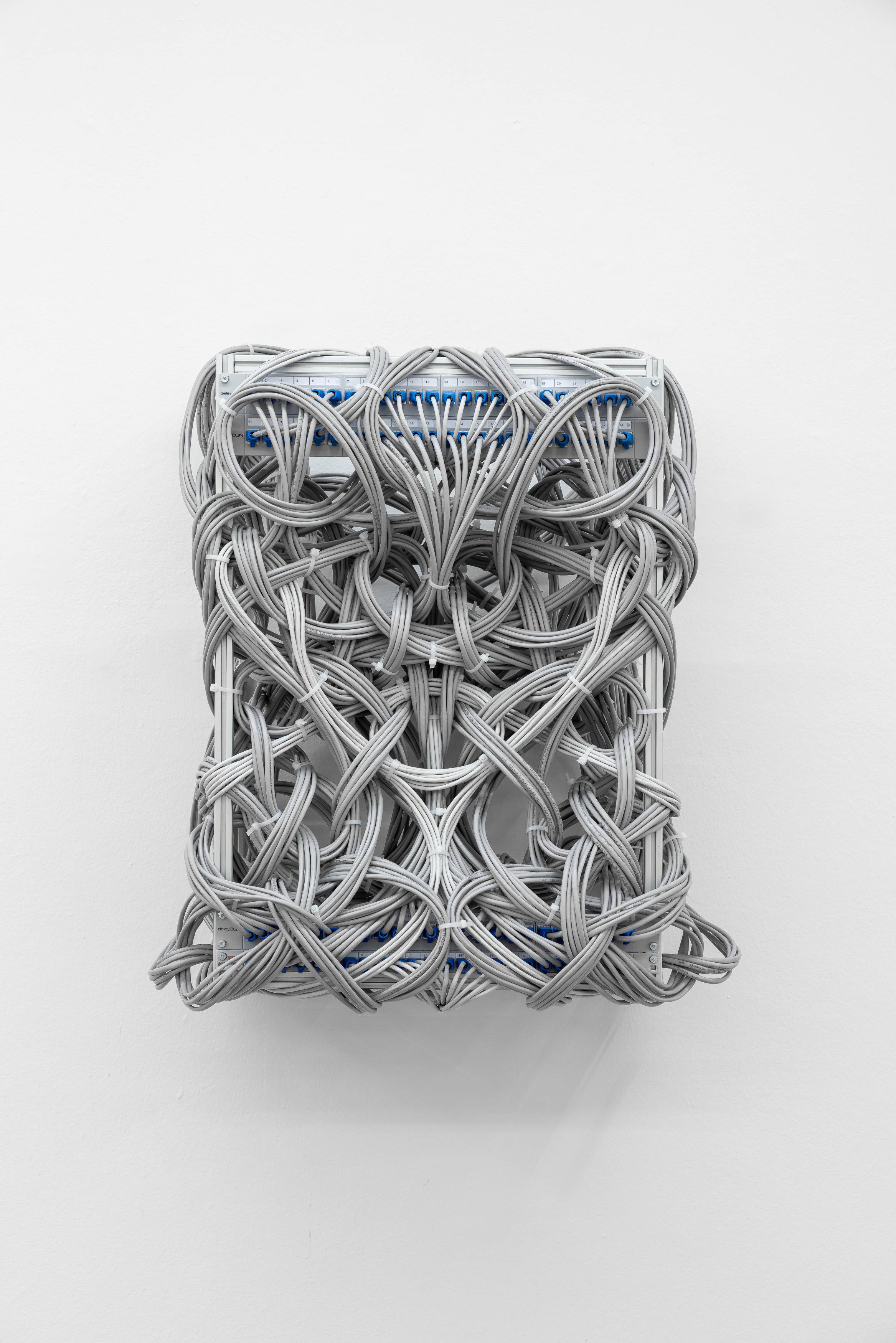 A wall-mounted sculpture of densely interwoven grey ethernet cables with blue connectors, forming a tight knotted rectangular mass against a white gallery wall